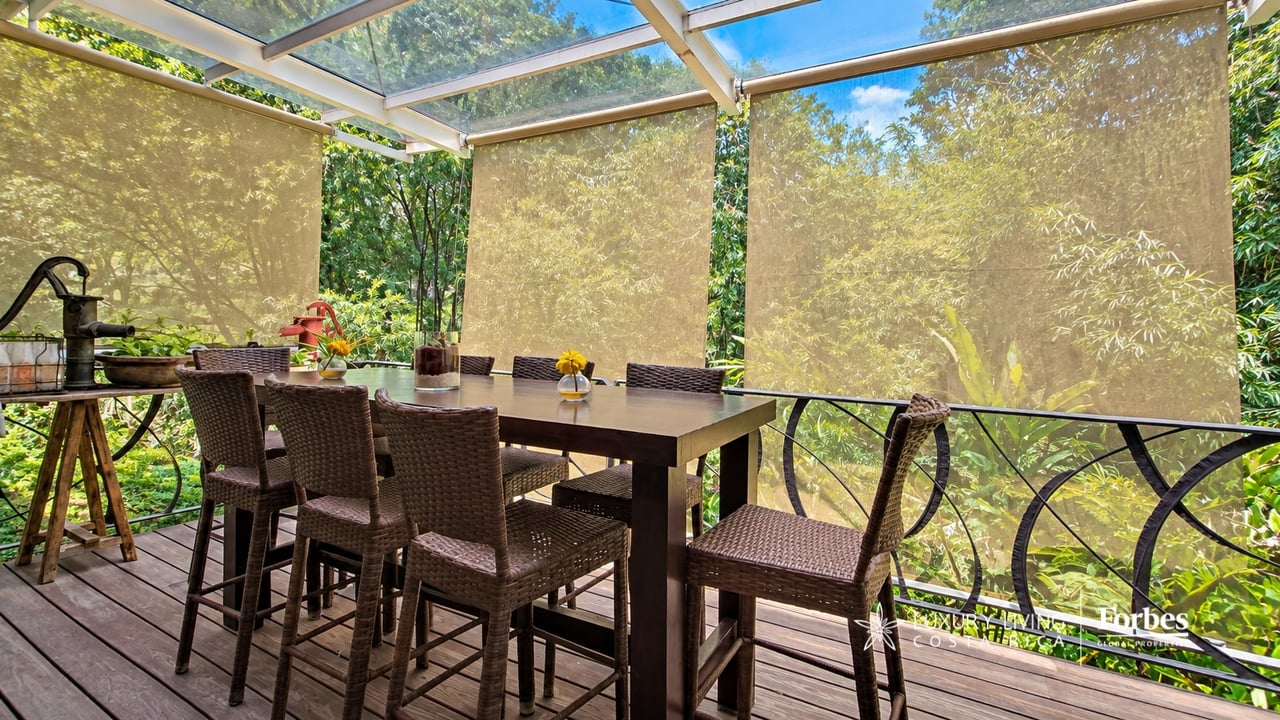 Covered terrace with a woven dining set, wood deck, and roll-down shades overlooking lush greenery.