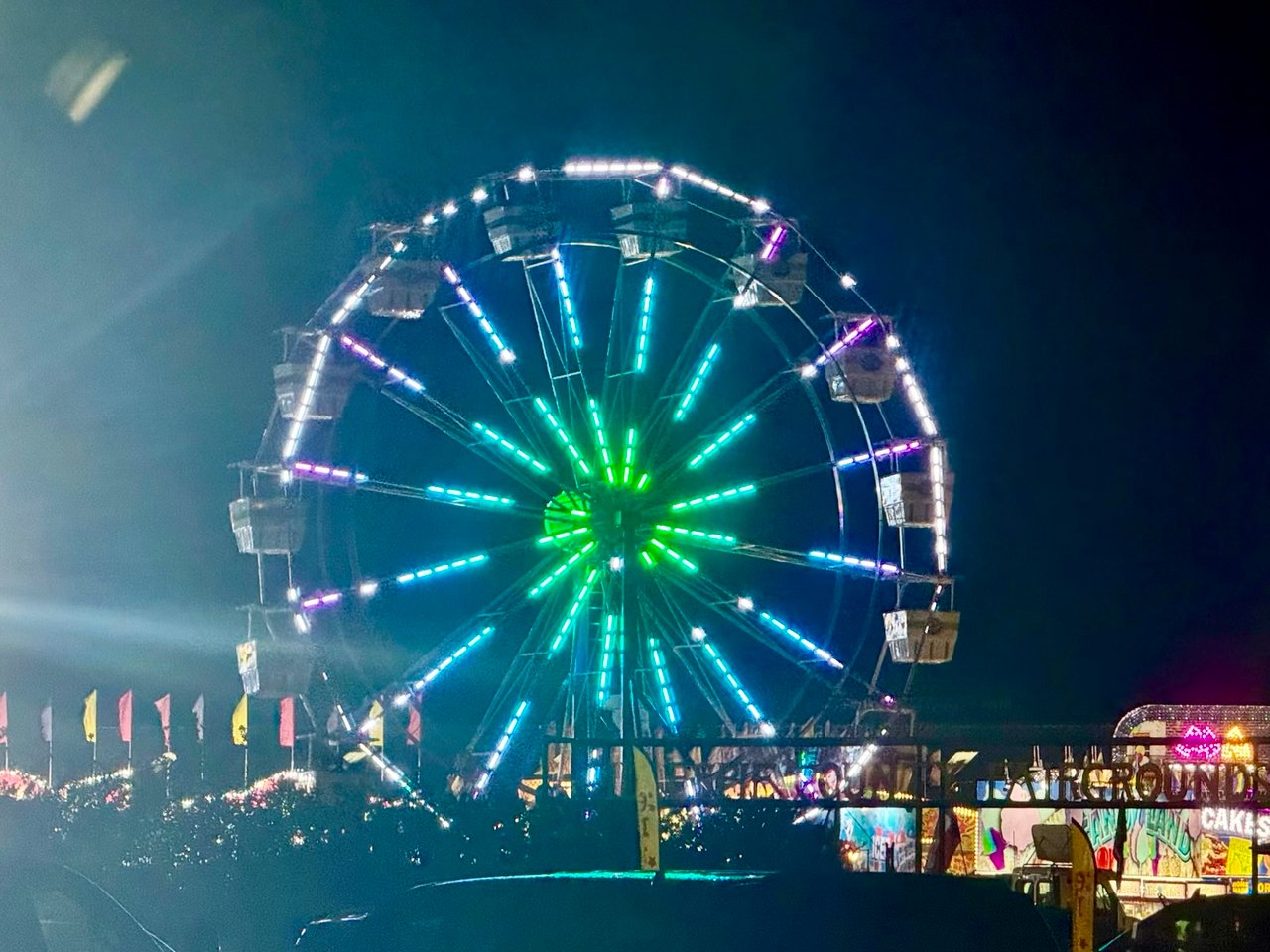 A neon-lit Ferris wheel towers over midway booths and flags at night.