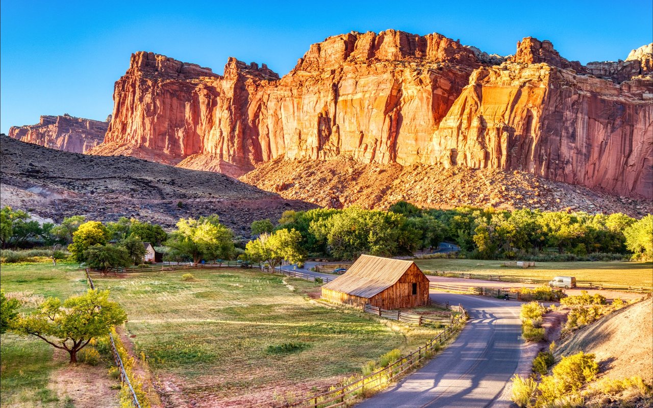 Red sandstone cliffs rise above green fields, a wooden barn, and a winding country road.