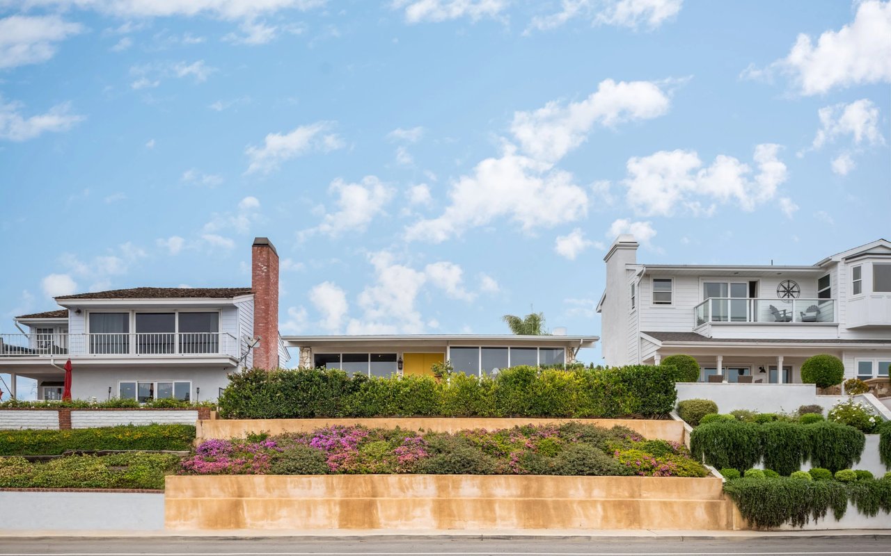 A row of houses on a hill, with lush green hedges and flowers in the foreground under a partly cloudy blue sky.