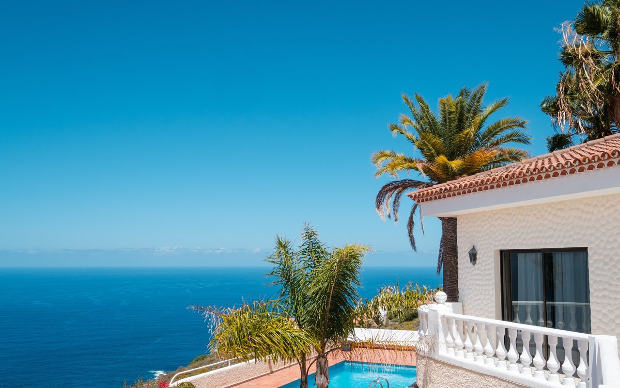 White coastal house with a tiled roof and balcony beside a pool overlooking the ocean.