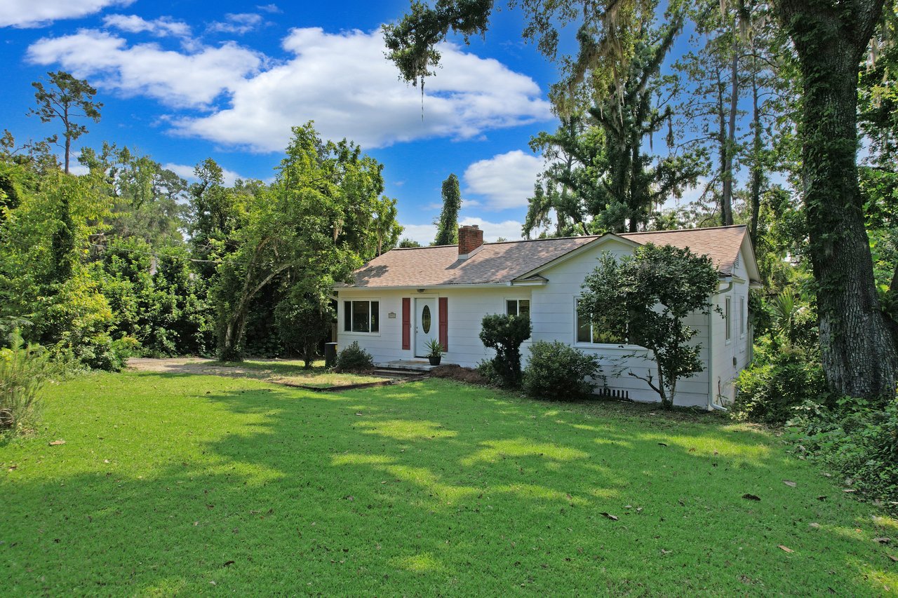 A cozy white house with a brown roof sits amid lush greenery. The front yard is expansive and sunlit under a bright blue sky with fluffy clouds.