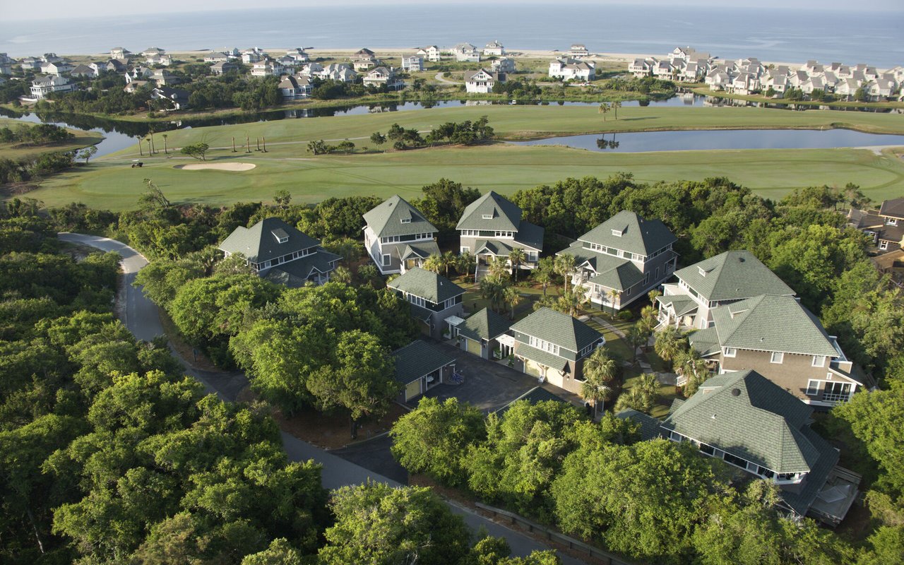 Aerial view of homes beside a golf course, with ponds and coastal water in the distance.