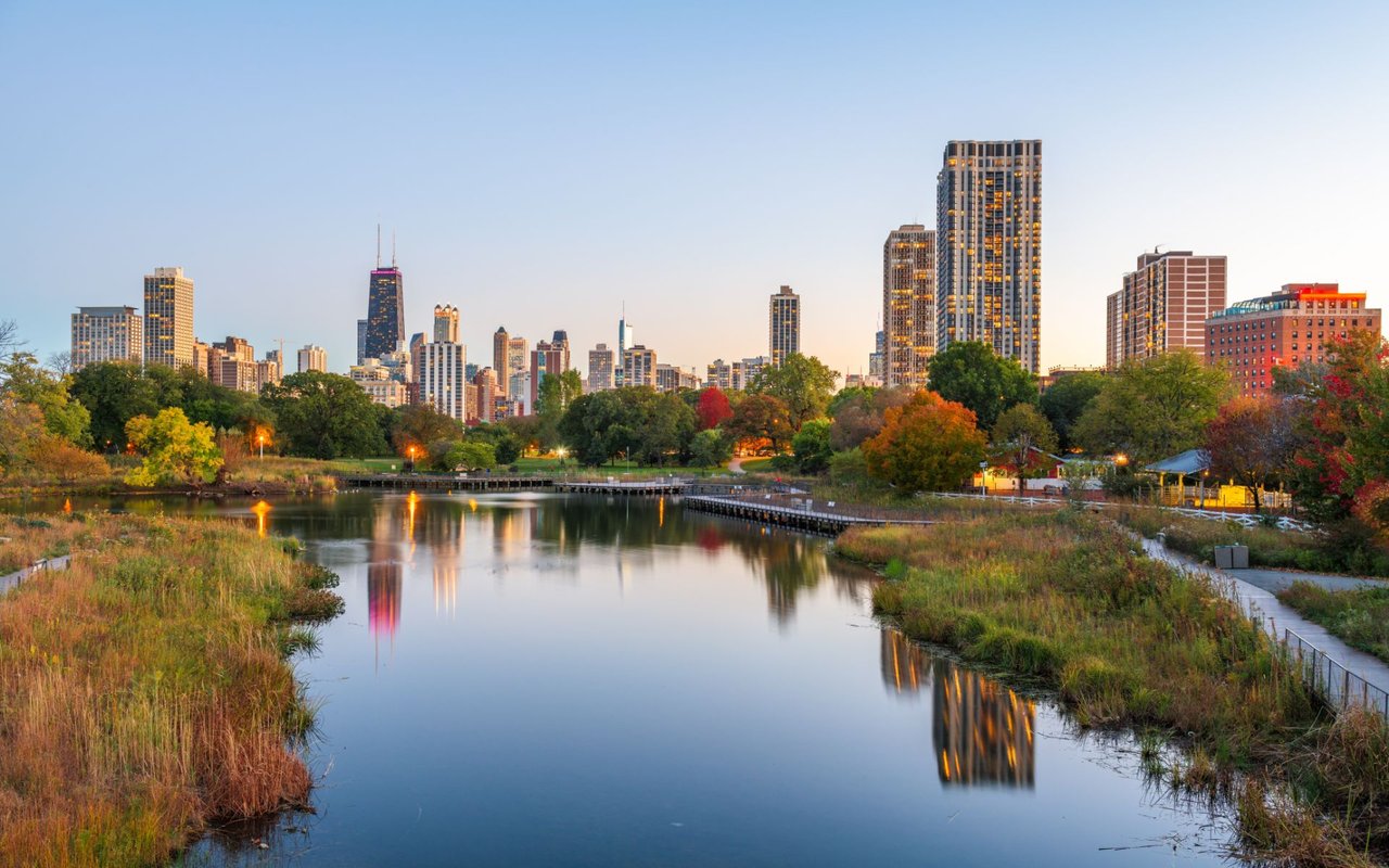 Chicago skyline reflected in a Lincoln Park pond, with autumn trees along the water.