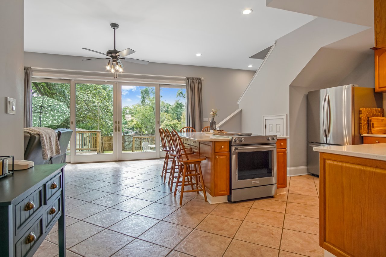 Spacious kitchen with tile floor, wooden cabinets, and a central island. Large windows overlook a green garden, creating a bright, inviting atmosphere.