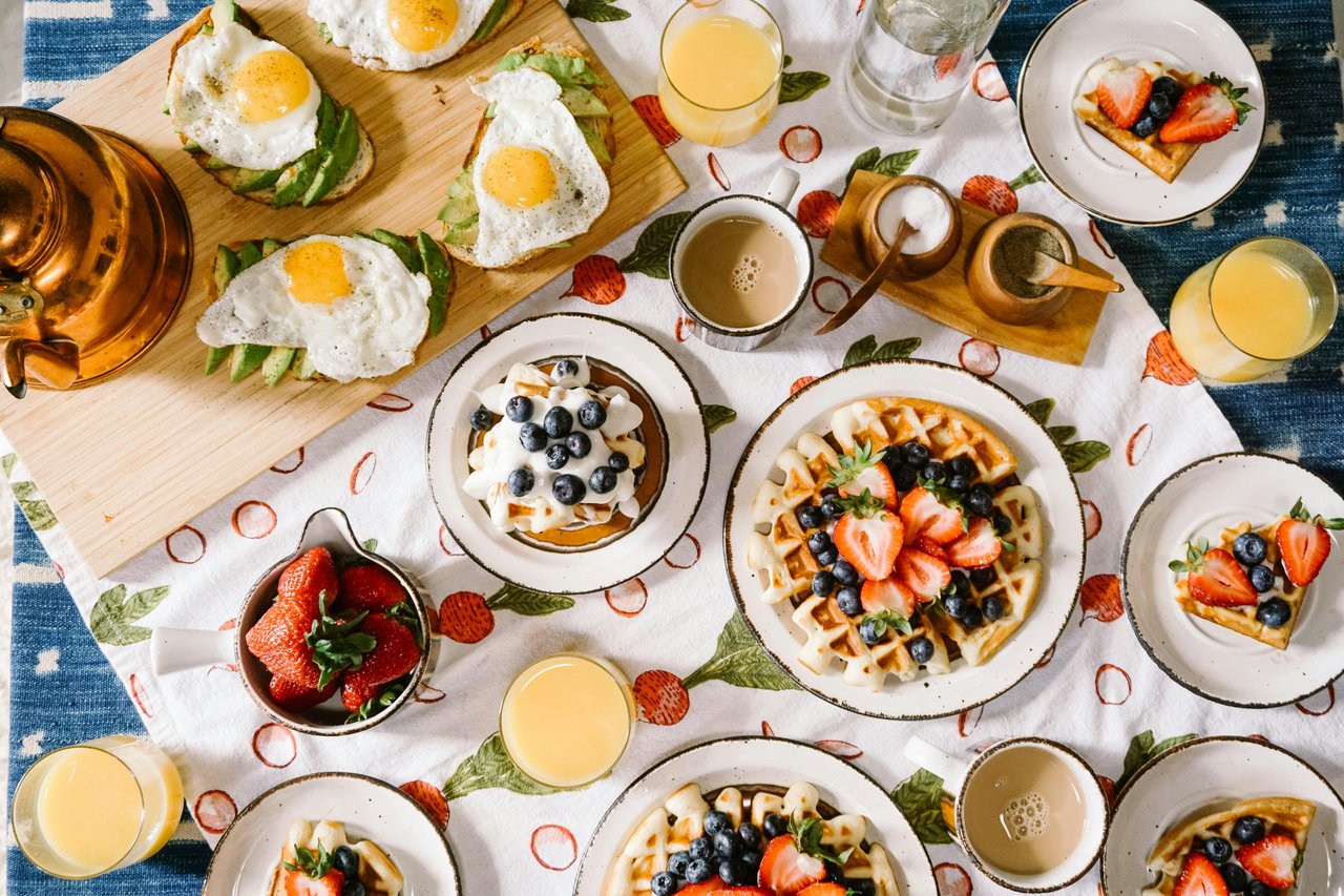 Breakfast spread with egg-topped avocado toast, berry waffles, pancakes, coffee, and orange juice.