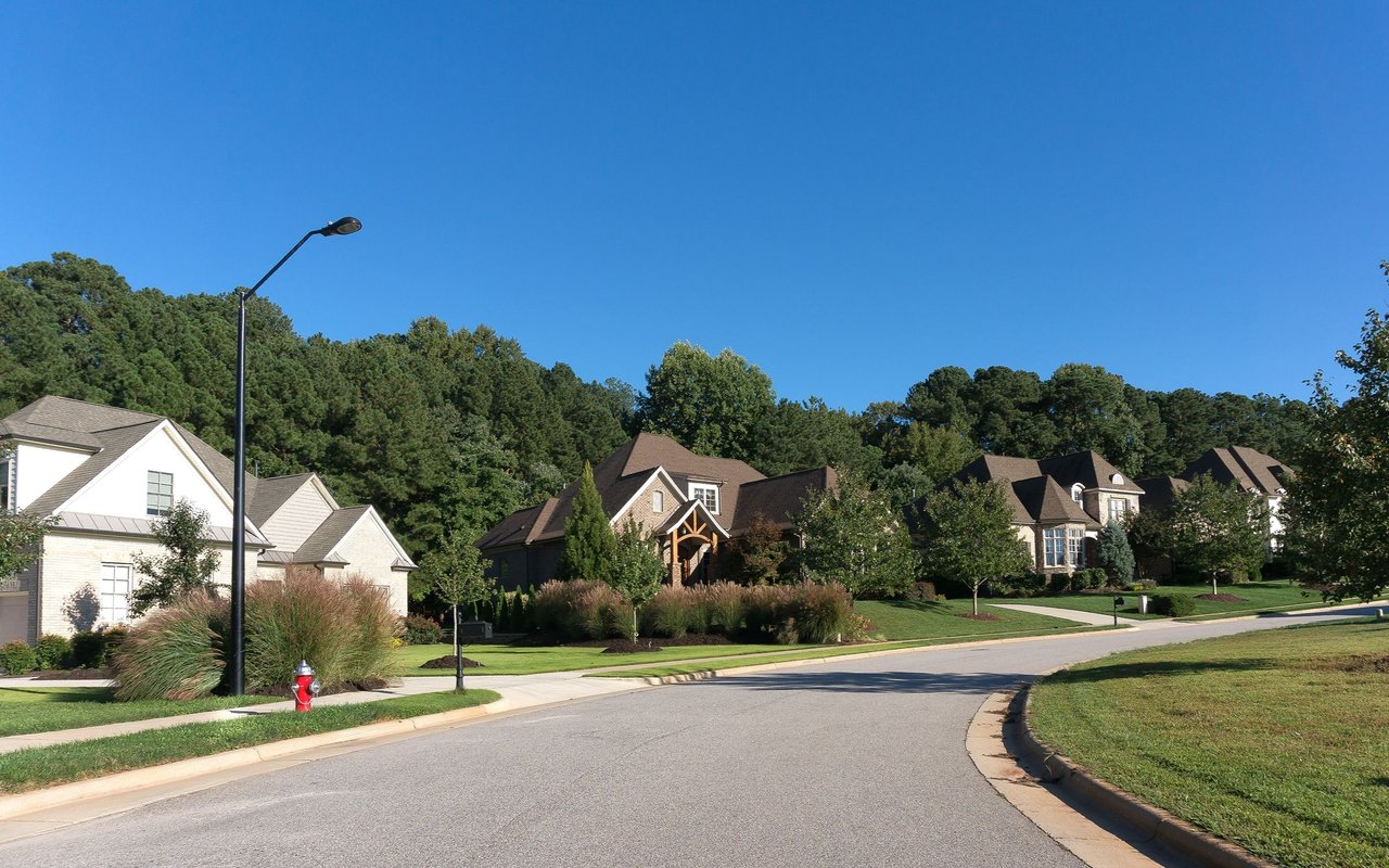 Residential street lined with detached homes and landscaped lawns beneath a clear blue sky.