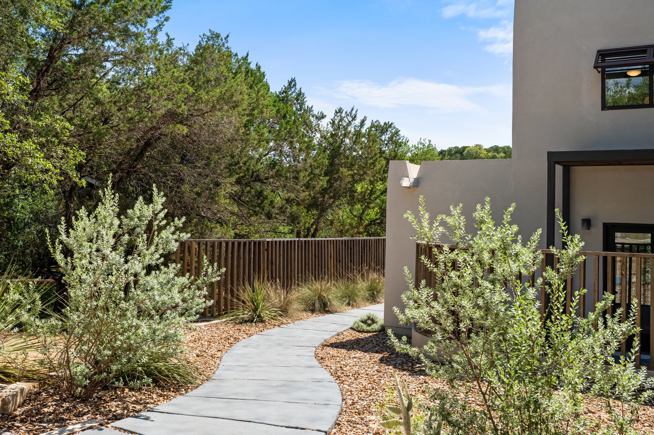 Modern stucco home beside a curved walkway bordered by rock mulch, shrubs, and mature trees.