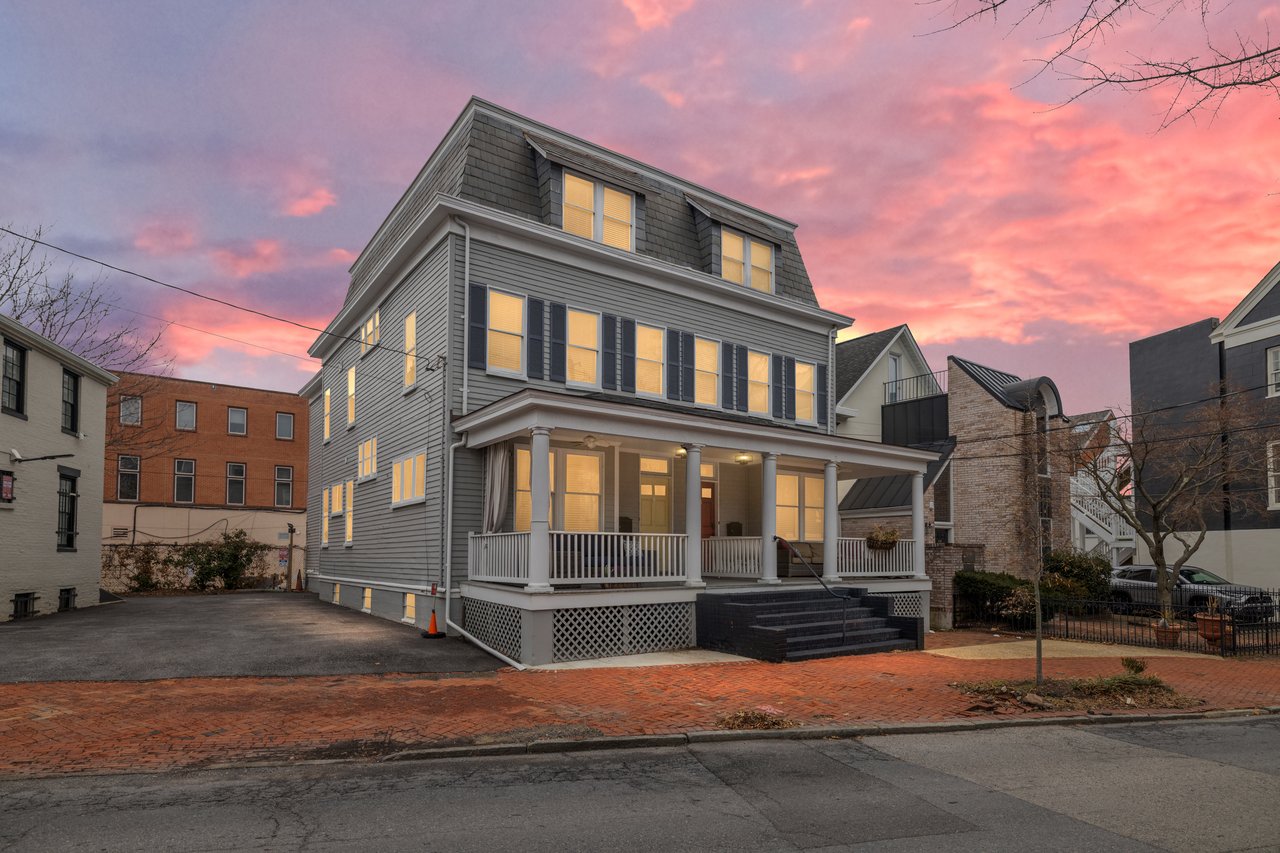 Gray multi-story house with a columned porch and glowing windows beneath a pink dusk sky.
