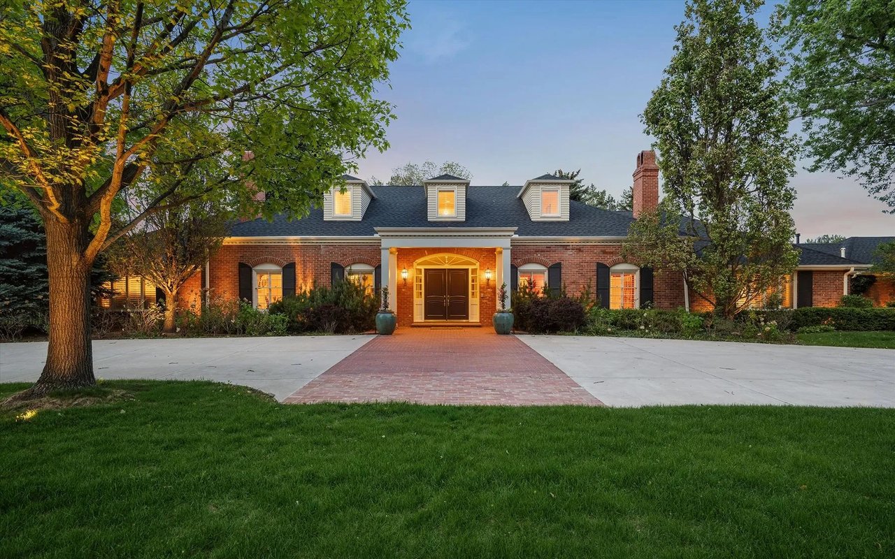 Red-brick house with three dormers, a warmly lit entrance, mature trees, and a broad driveway.