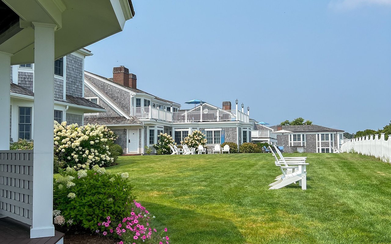 Shingle-style coastal home beyond a lawn, with white Adirondack chairs and flowering hydrangeas.
