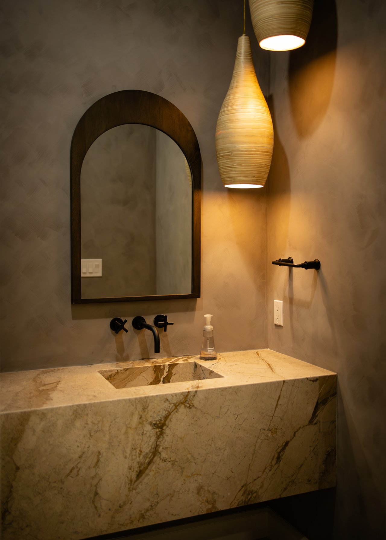Bathroom vanity with a veined marble sink, black wall-mounted faucet, arched mirror, and pendant lights.