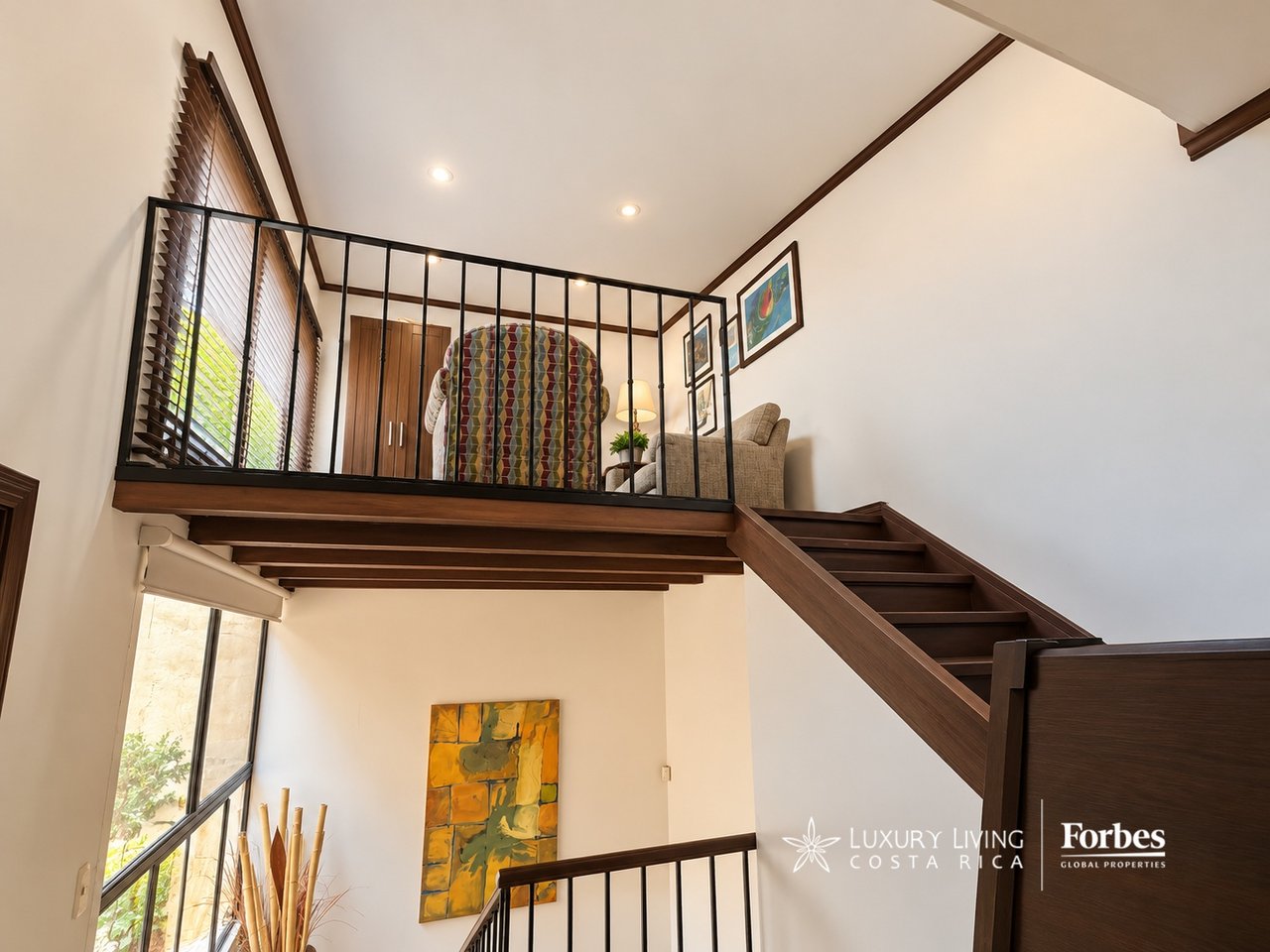 Wooden staircase rises to a furnished mezzanine with a black metal railing and tall windows.
