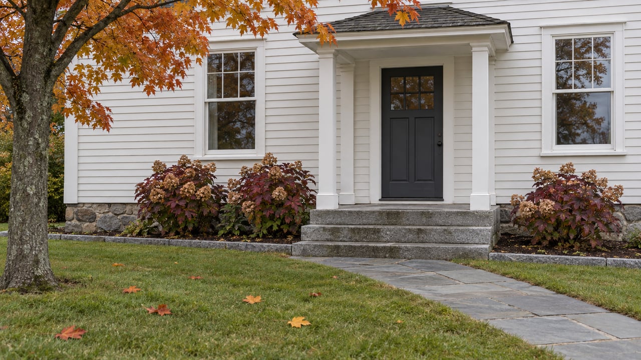 White clapboard house with a charcoal front door, granite steps, bluestone approach and amber maple foliage above an autumn lawn.