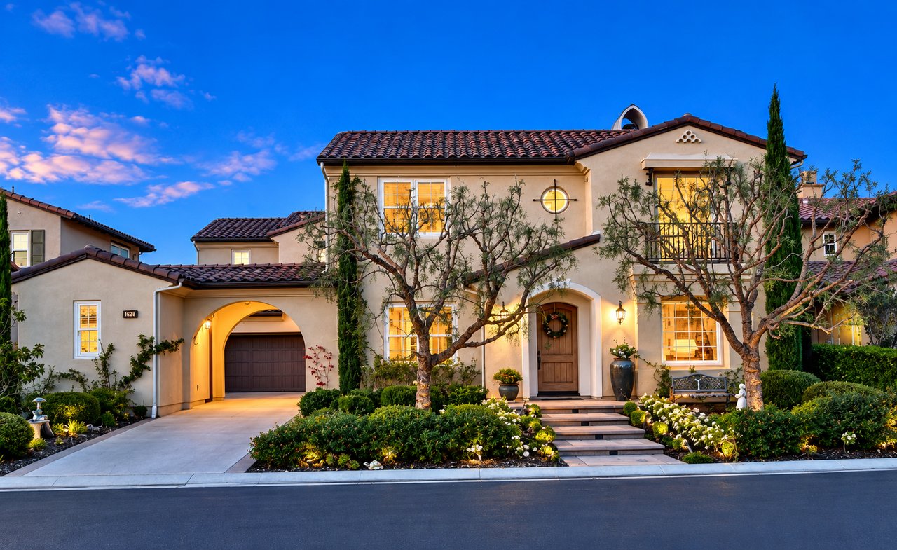 Two-story stucco home with a tiled roof, arched garage entrance, and warmly lit windows at dusk.