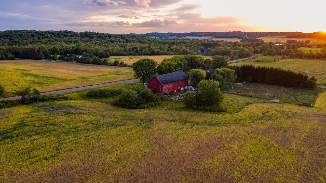 Aerial view of rolling farmland and wooded hills, with a red barn in the foreground at sunset.
