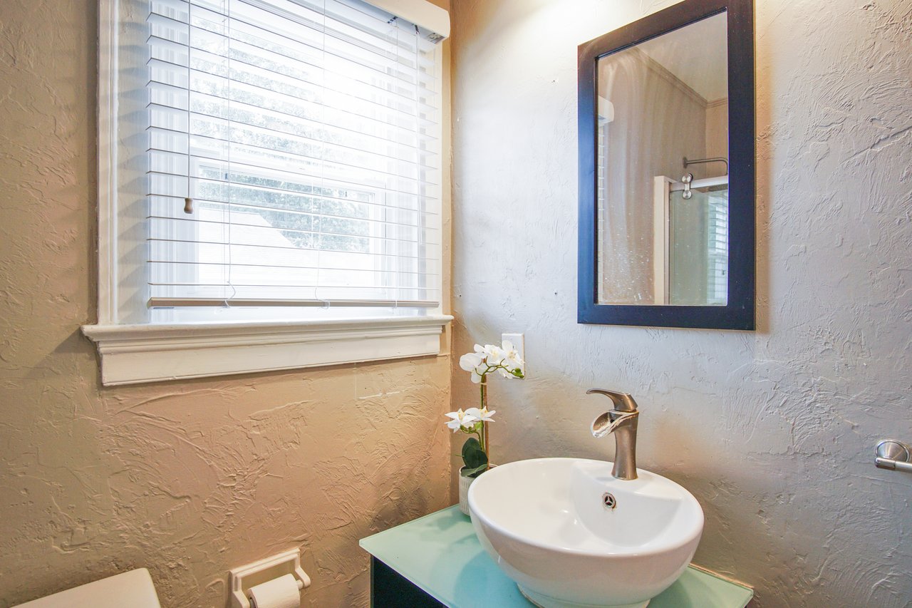 A cozy bathroom with textured beige walls, a white vessel sink, sleek faucet, small orchid plant, and a window with blinds, conveying a serene ambiance.