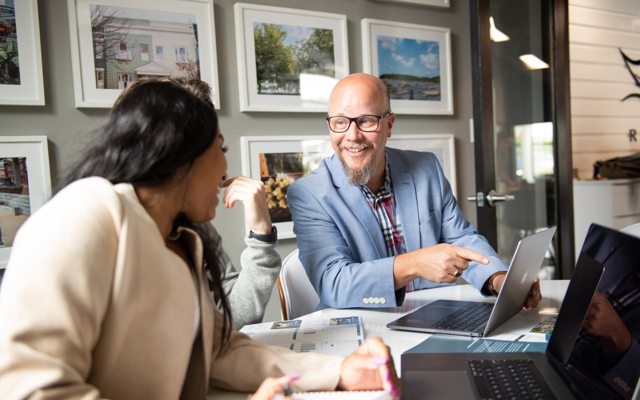Real estate agent discusses pricing with clients around a table with laptops and property brochures.