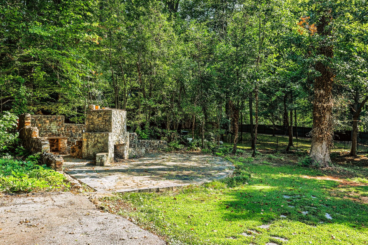 Wooded backyard patio with a built-in stone fireplace and mature trees.