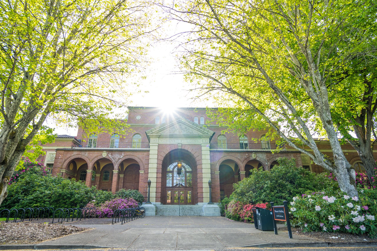 Brick Women's Building with arched entrance, flowering shrubs, and leafy trees at Oregon State University.