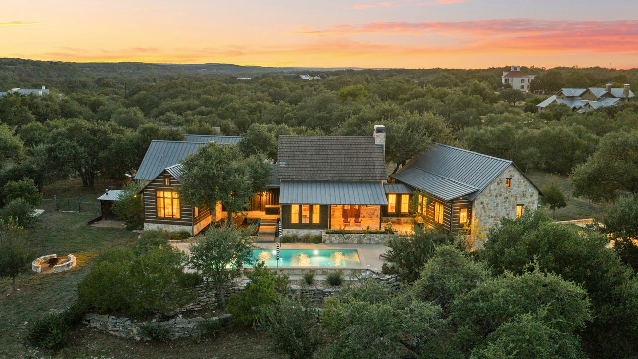 Aerial view of a stone-and-wood home with a lit swimming pool among wooded hills at sunset.