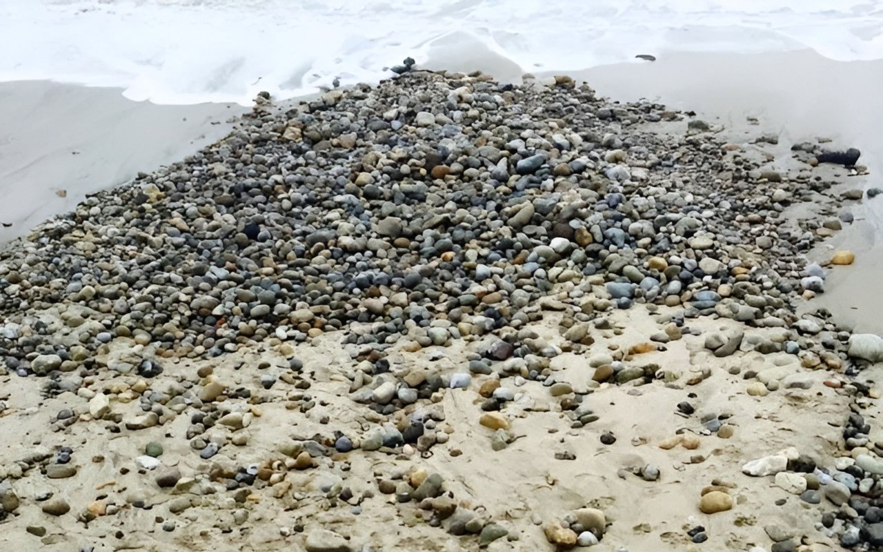 Smooth pebbles cover a sandy beach as white surf reaches the shore.