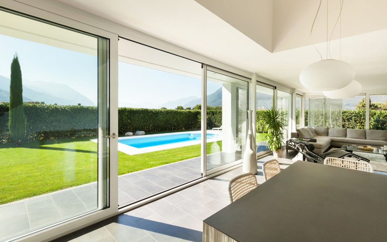 A luxurious modern living area with a large dining table and glass doors leading to a backyard with a pool and scenic views.