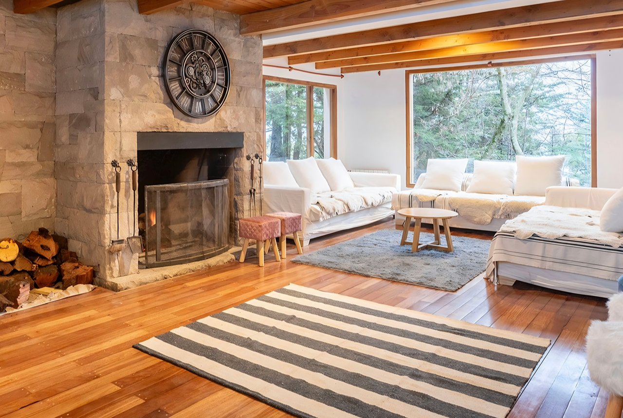 Living room with a stone fireplace, exposed beams, and large windows overlooking trees.