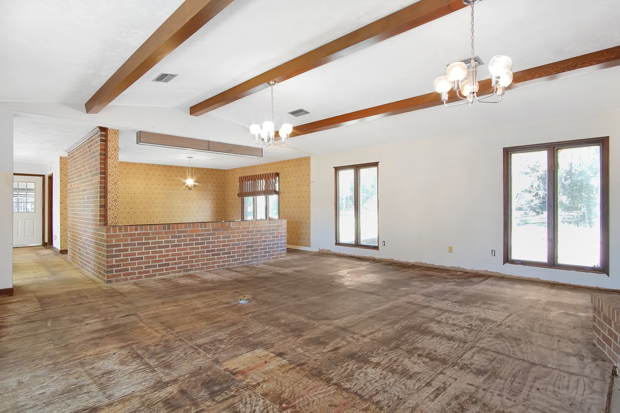 Spacious room with exposed wooden beams, brick accent walls, and vintage wallpaper. Large windows provide natural light. Floor appears unfinished.