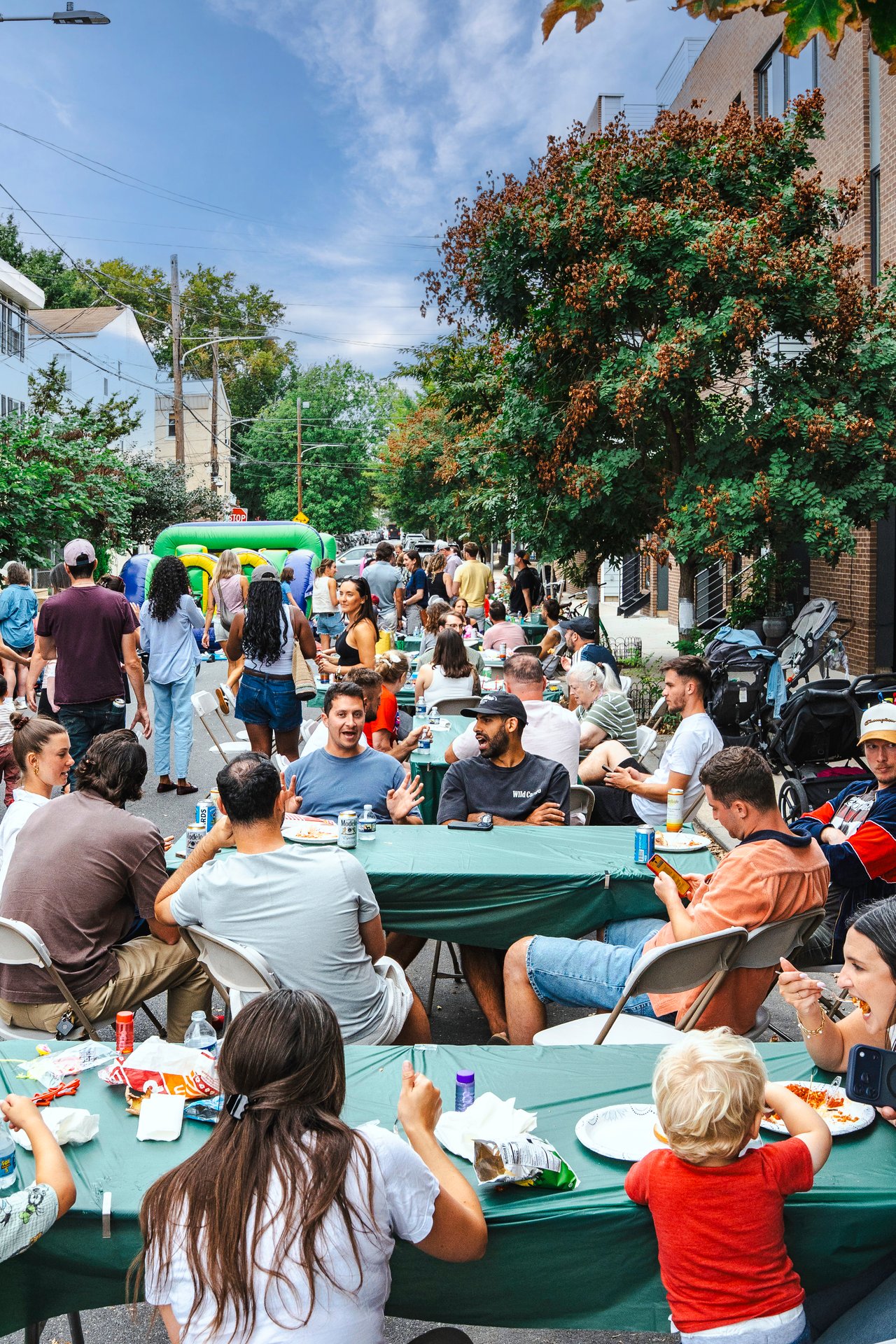 Neighbors share a meal at long tables during a street block party, with an inflatable play structure nearby.