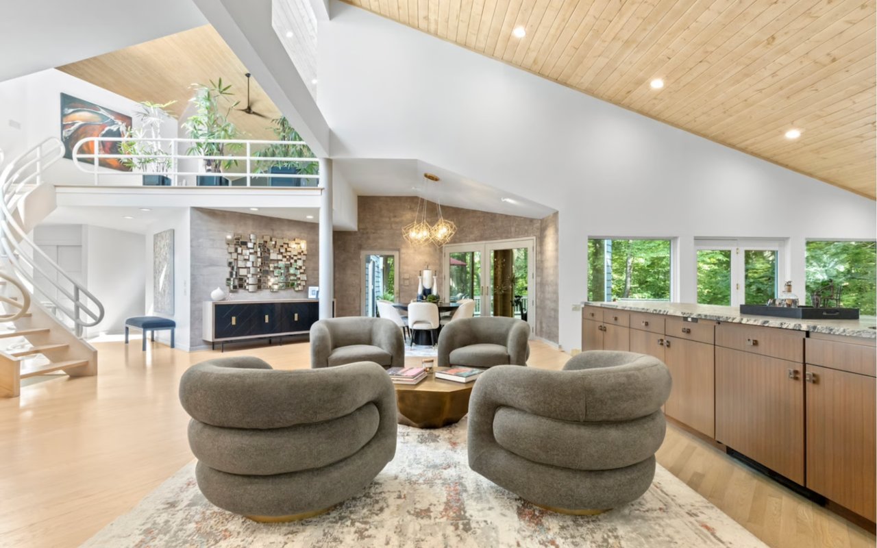 Open-plan living room with sculptural gray chairs, a curved staircase, and a vaulted wood ceiling.