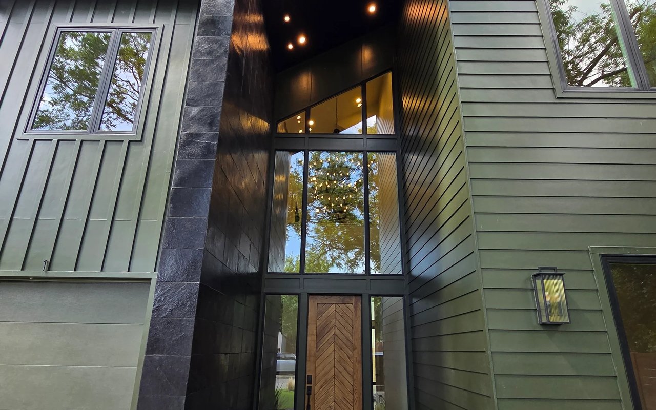 Modern home entrance with a chevron-patterned wood door, tall windows, and green siding.