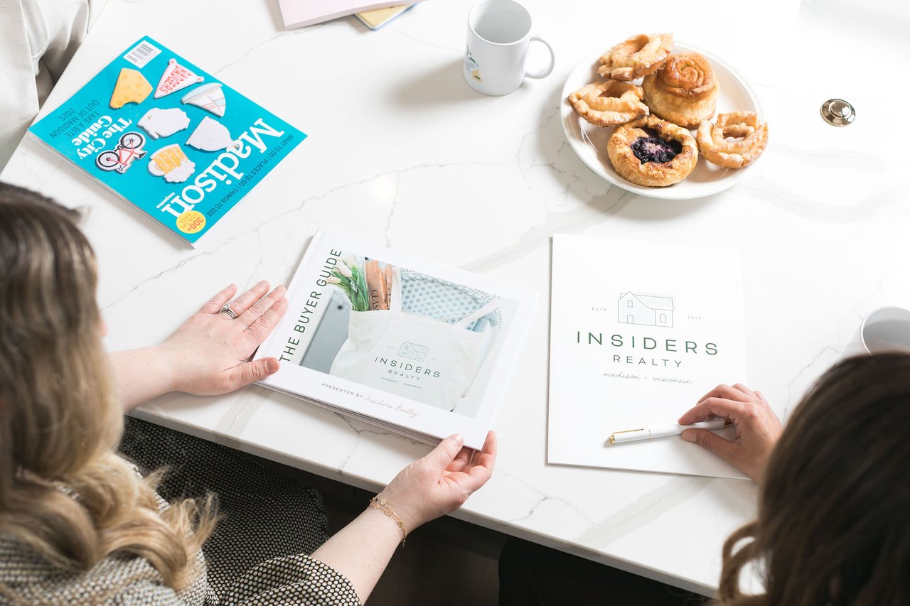 Two women sitting at a white marble table reviewing an Insiders Realty Buyer Guide and Madison Magazine, illustrating the expert planning required to navigate the $500,000 to $800,000 price range in the 2026 Madison, Wisconsin real estate market.