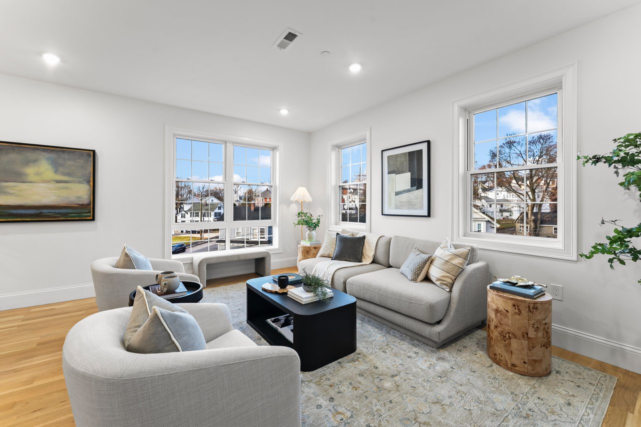 Staged living room with gray sofas, a black coffee table, and large windows overlooking neighboring homes.