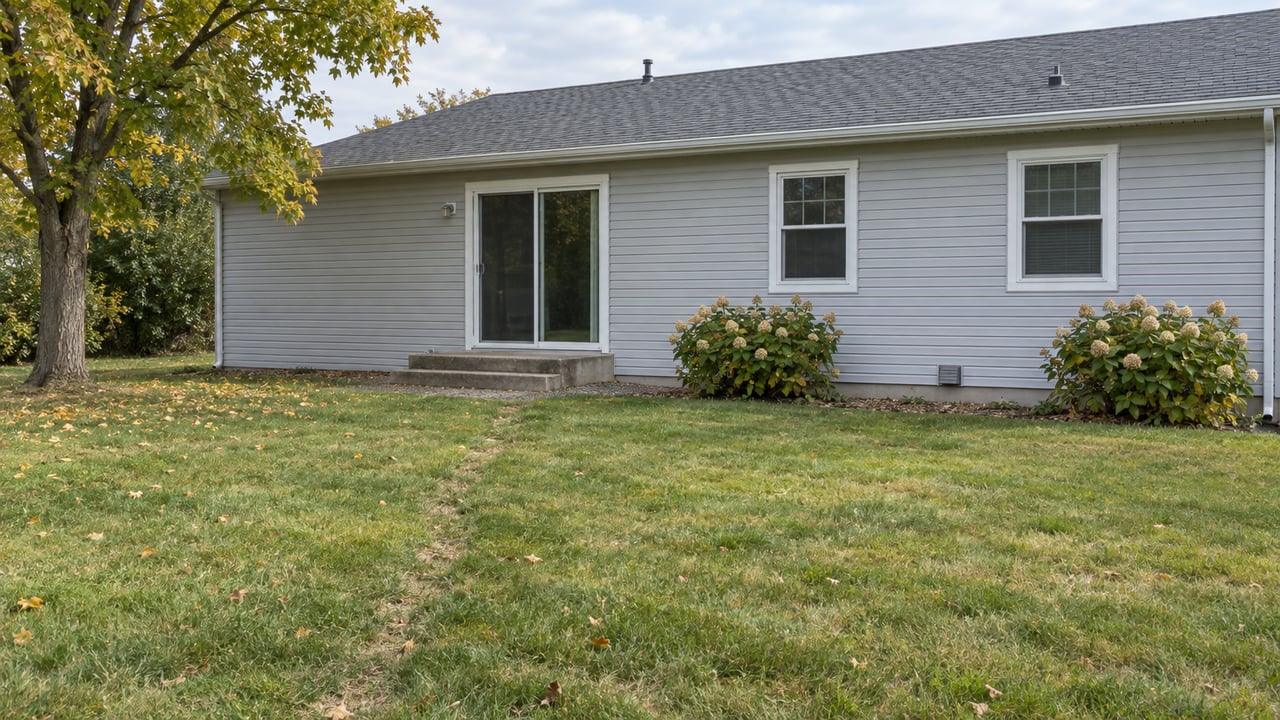 Modest ranch house with gray siding, sliding glass door, and two steps beyond a broad, slightly straw-toned lawn.