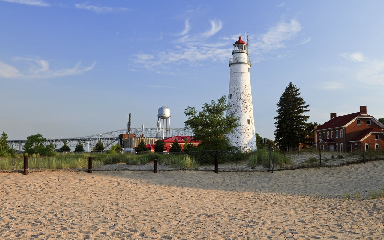 Fort Gratiot Lighthouse beside a sandy beach, with a bridge and brick buildings in the background.