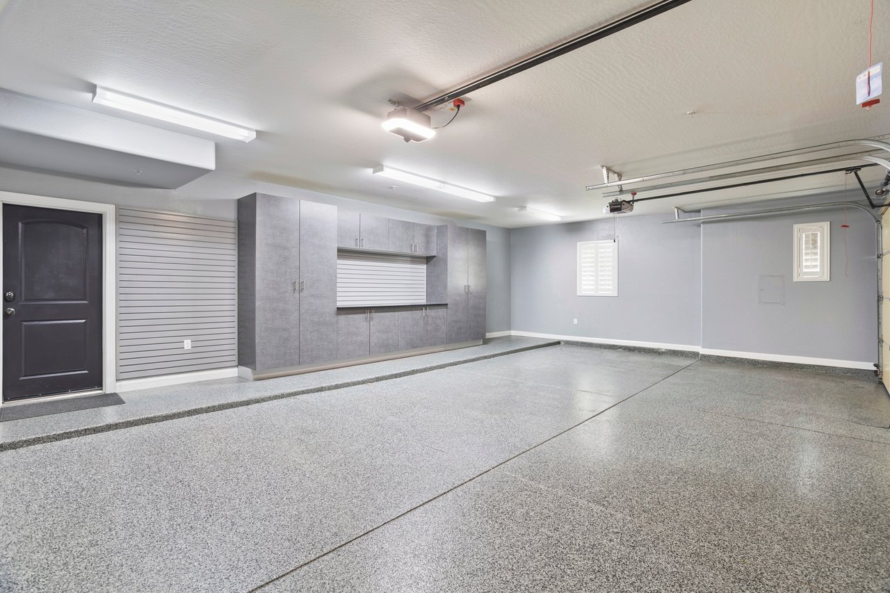 Spacious finished garage with speckled epoxy floor, gray built-in cabinets, overhead fluorescent lights, and garage door opener rails.