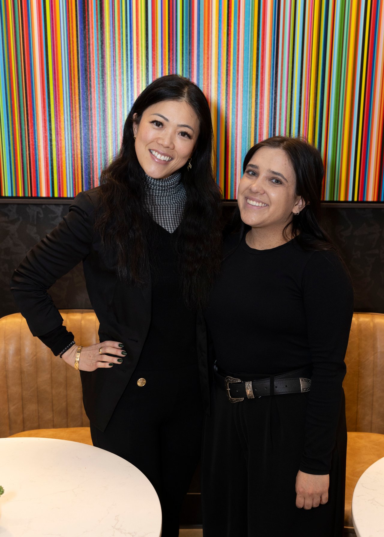 Two women pose together in black outfits in front of colorful striped artwork.