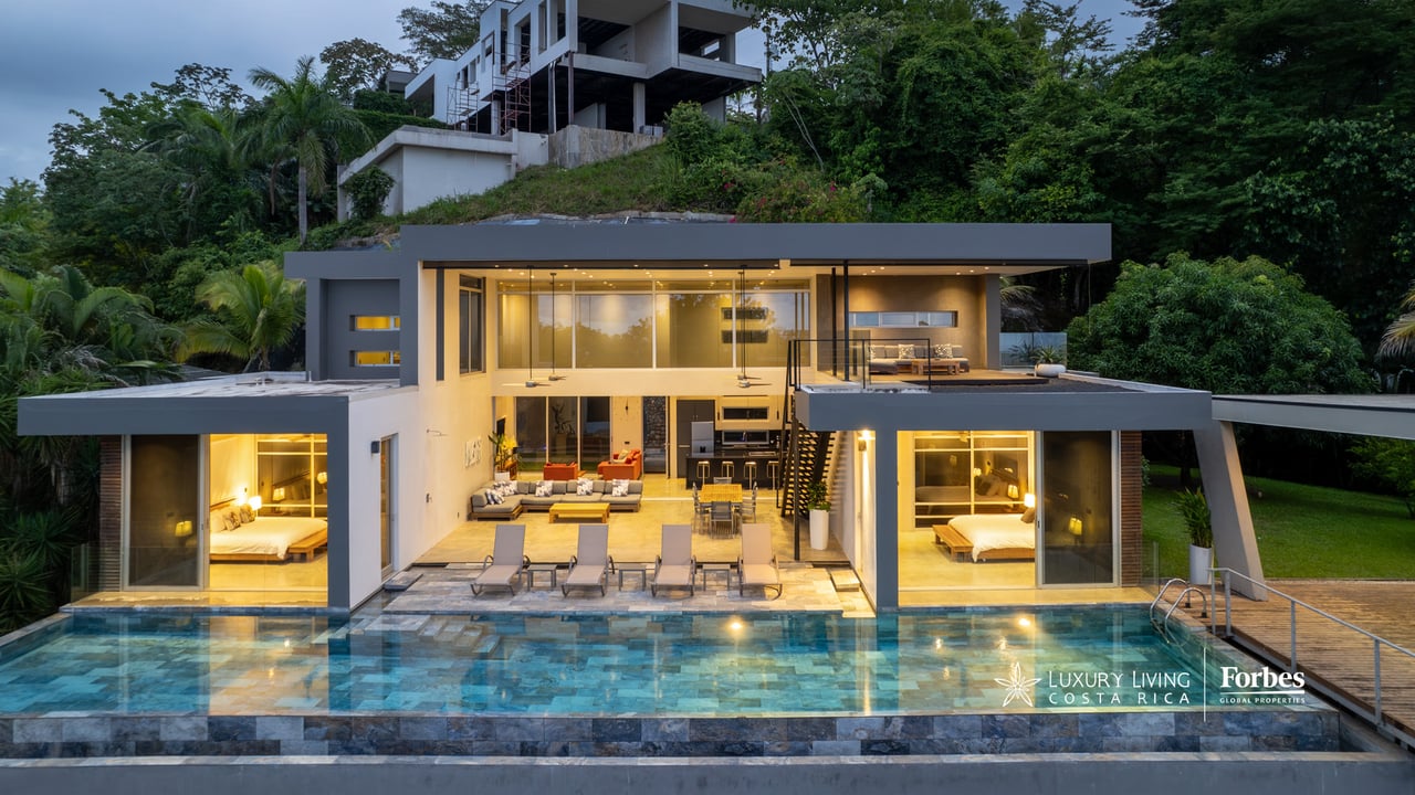 Modern hillside home with glass walls and an illuminated pool at dusk.
