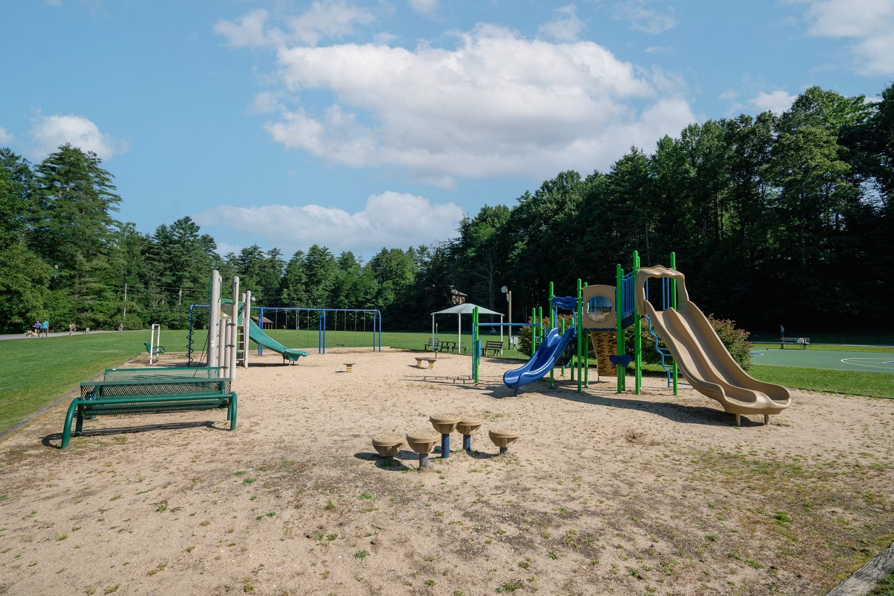 A playground with slides, climbing structures, swings, and seating benches on a sandy surface, surrounded by grassy fields and trees under a partly cloudy sky—an ideal platform for families to relax and spend time together.