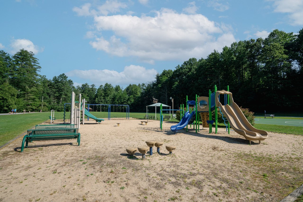 Alt=A playground with slides, climbing structures, swings, and seating benches on a sandy surface, surrounded by grassy fields and trees under a partly cloudy sky—an ideal platform for families to relax and spend time together.