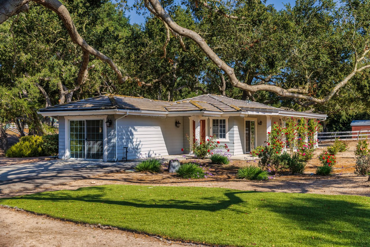 Single-story white house beneath sprawling oak trees, with red roses and a green front lawn.