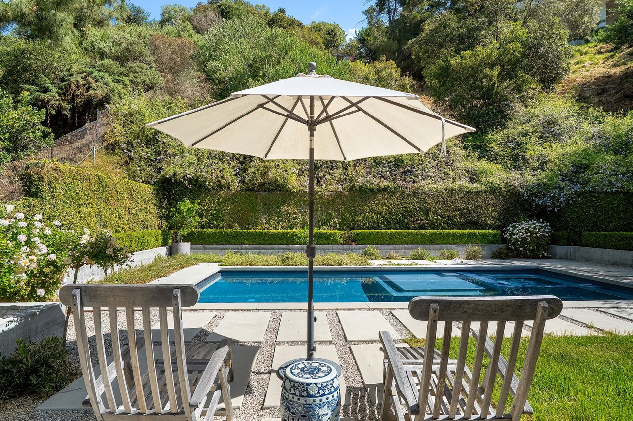 Swimming pool framed by patio chairs, a white umbrella, and lush flowering hedges.