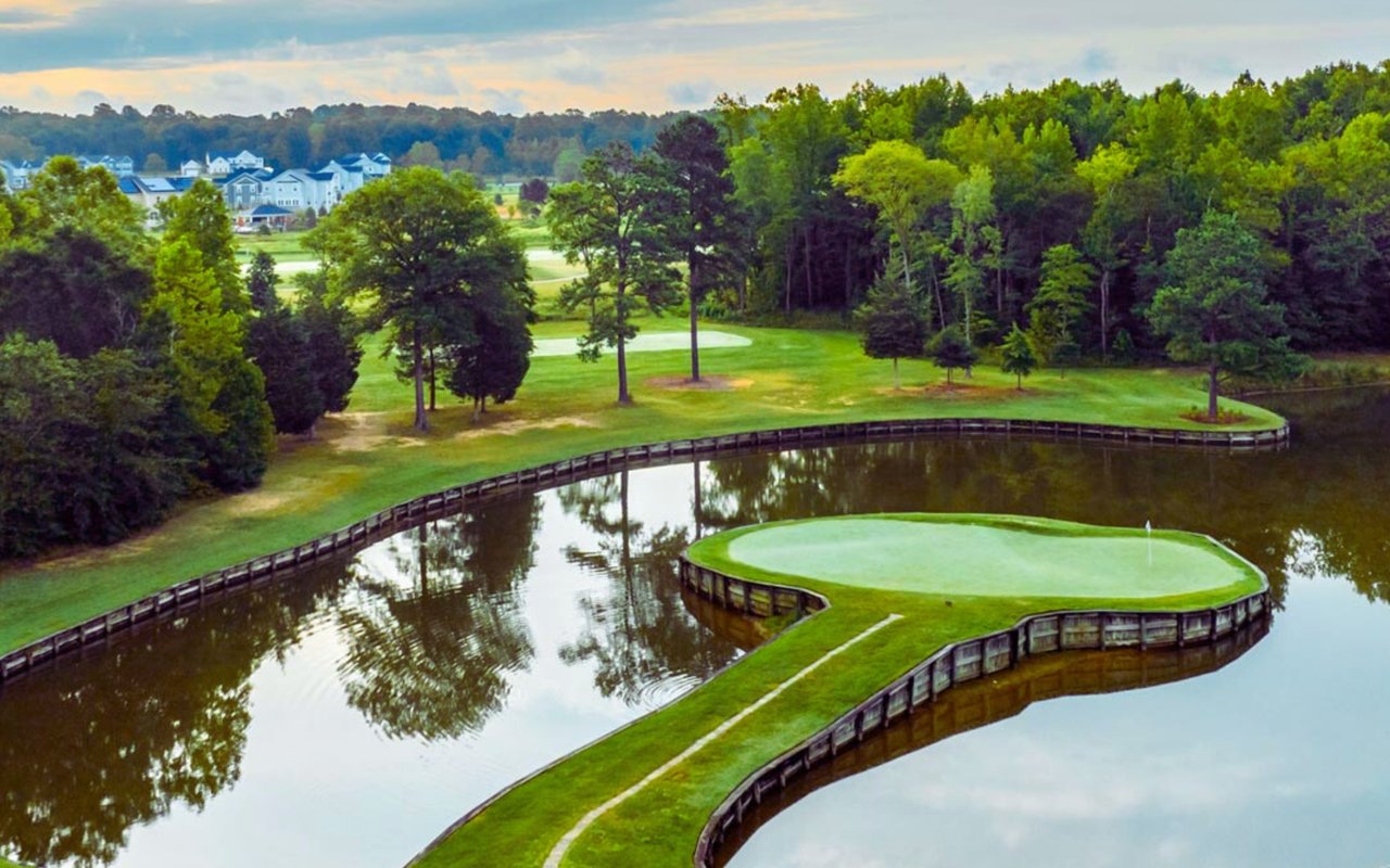 Golf course green surrounded by water, with a curving wooden retaining wall and wooded grounds.