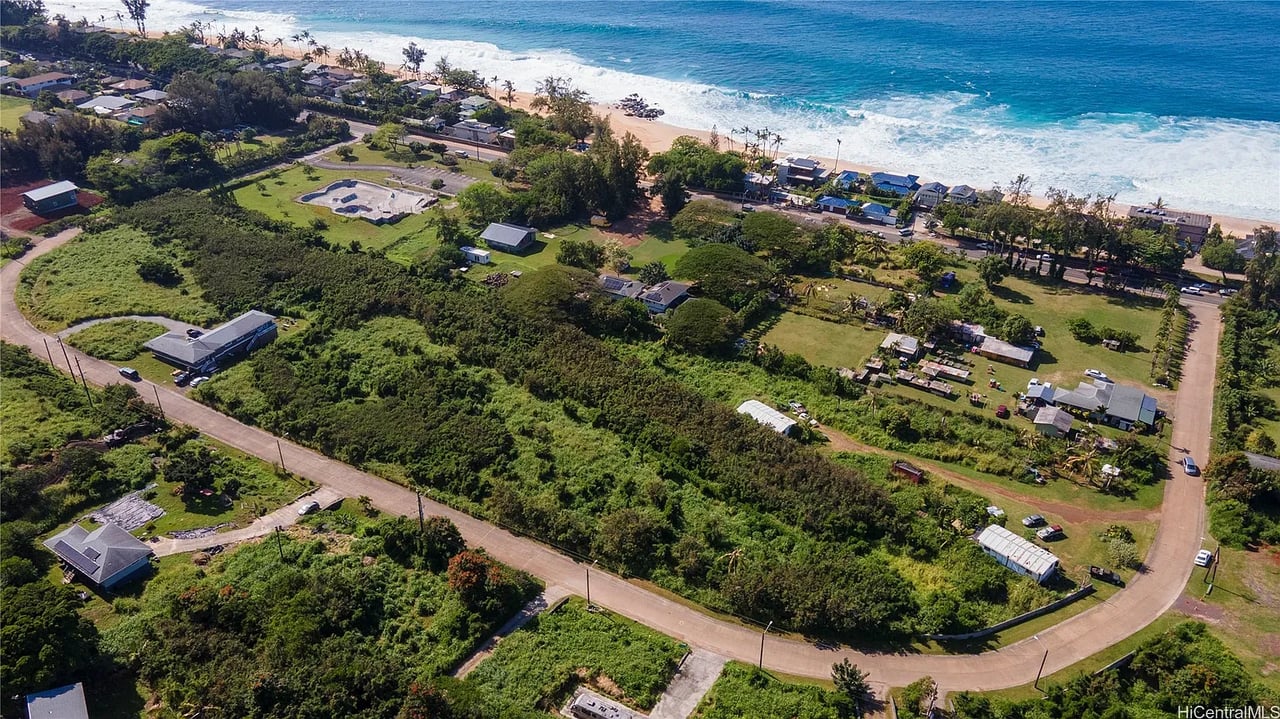 Aerial view of a green coastal parcel, neighborhood homes, sandy beach, and turquoise ocean.