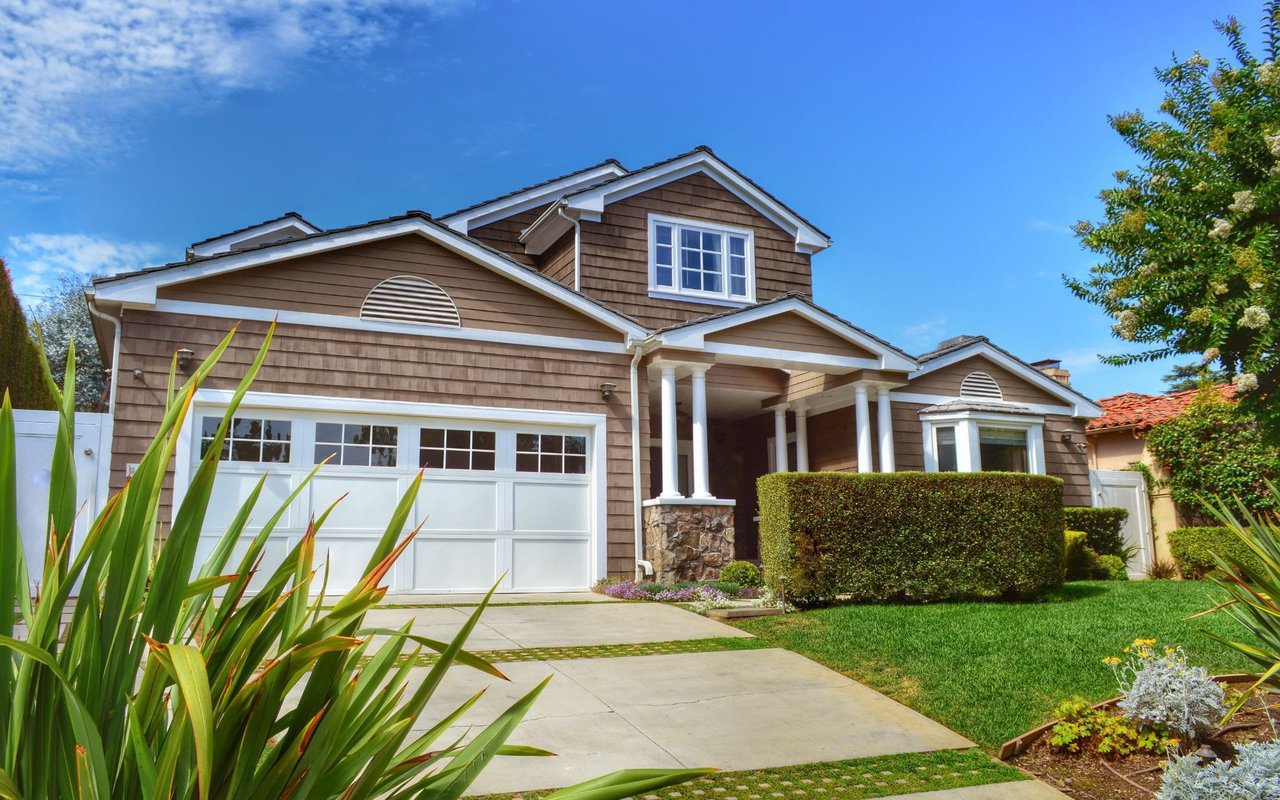Lush green landscaping, including a neatly trimmed hedge and various plants, frames the house. 
