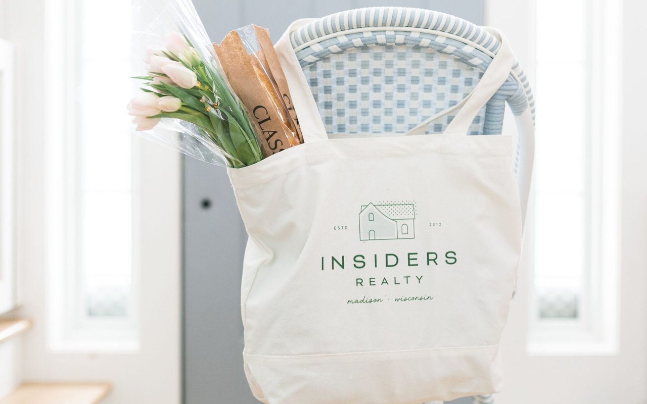 A canvas tote bag with the Insider Realty logo rests on a metal chair, its top filled with flowers and bread.