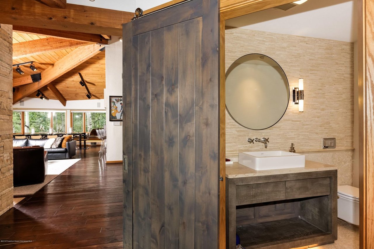 Bathroom vanity with a vessel sink and round mirror; an open barn door reveals a timber-beamed living room.