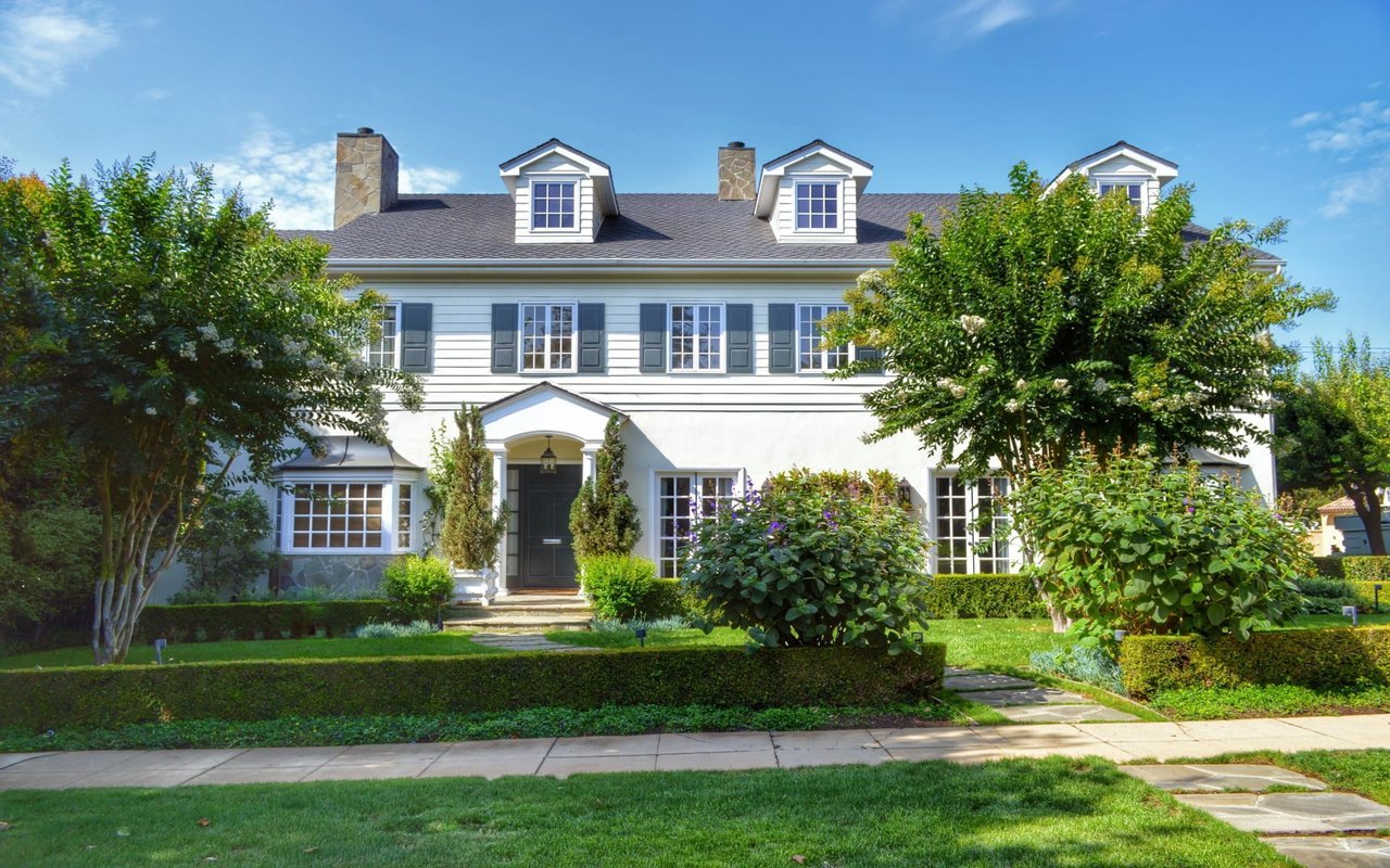 A large Colonial-style house with many windows, a front lawn, a walkway to the front door, and surrounded by greeneries.