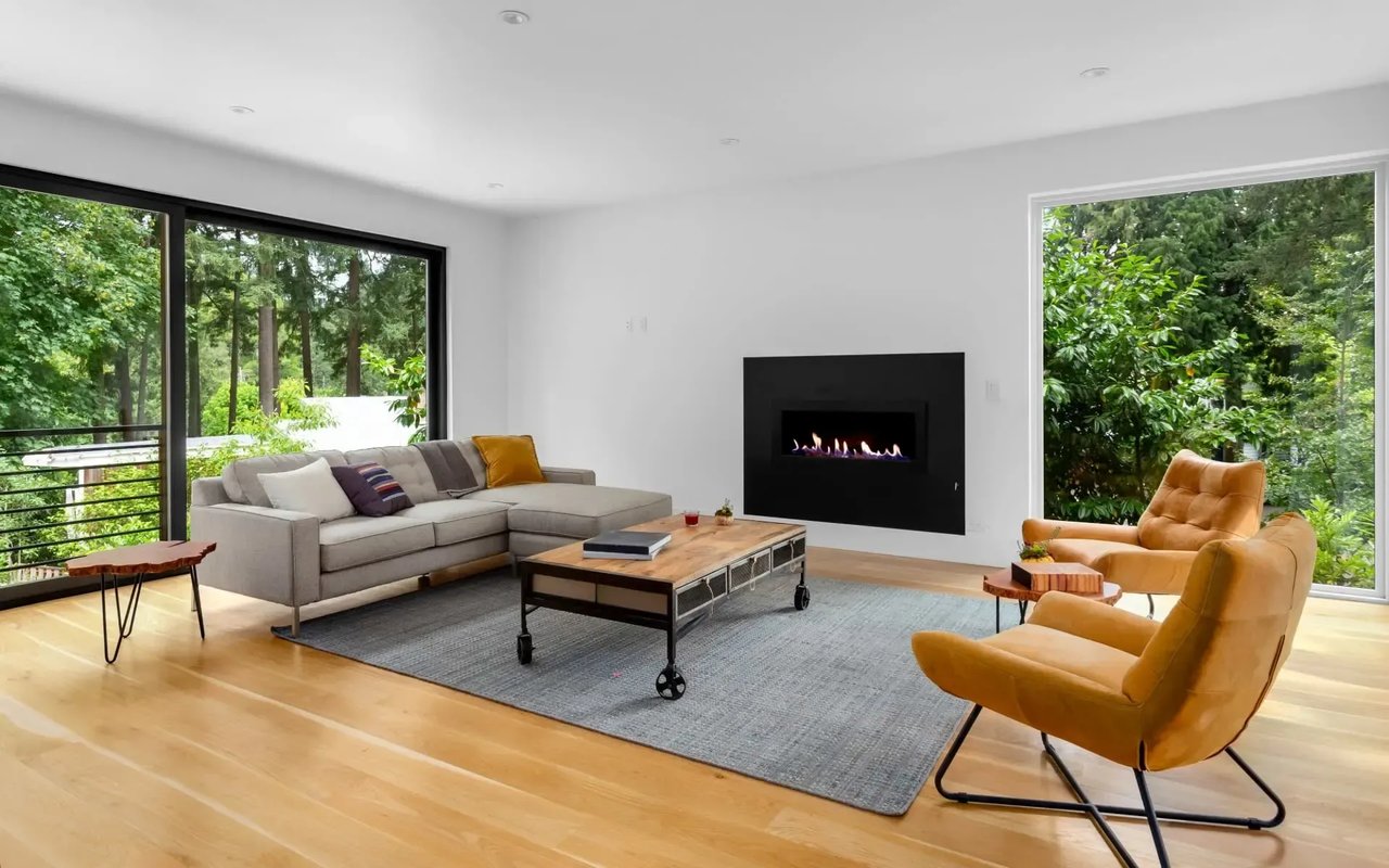 A living room with large windows overlooking a forested area, featuring a sectional sofa, a coffee table, and a fireplace.